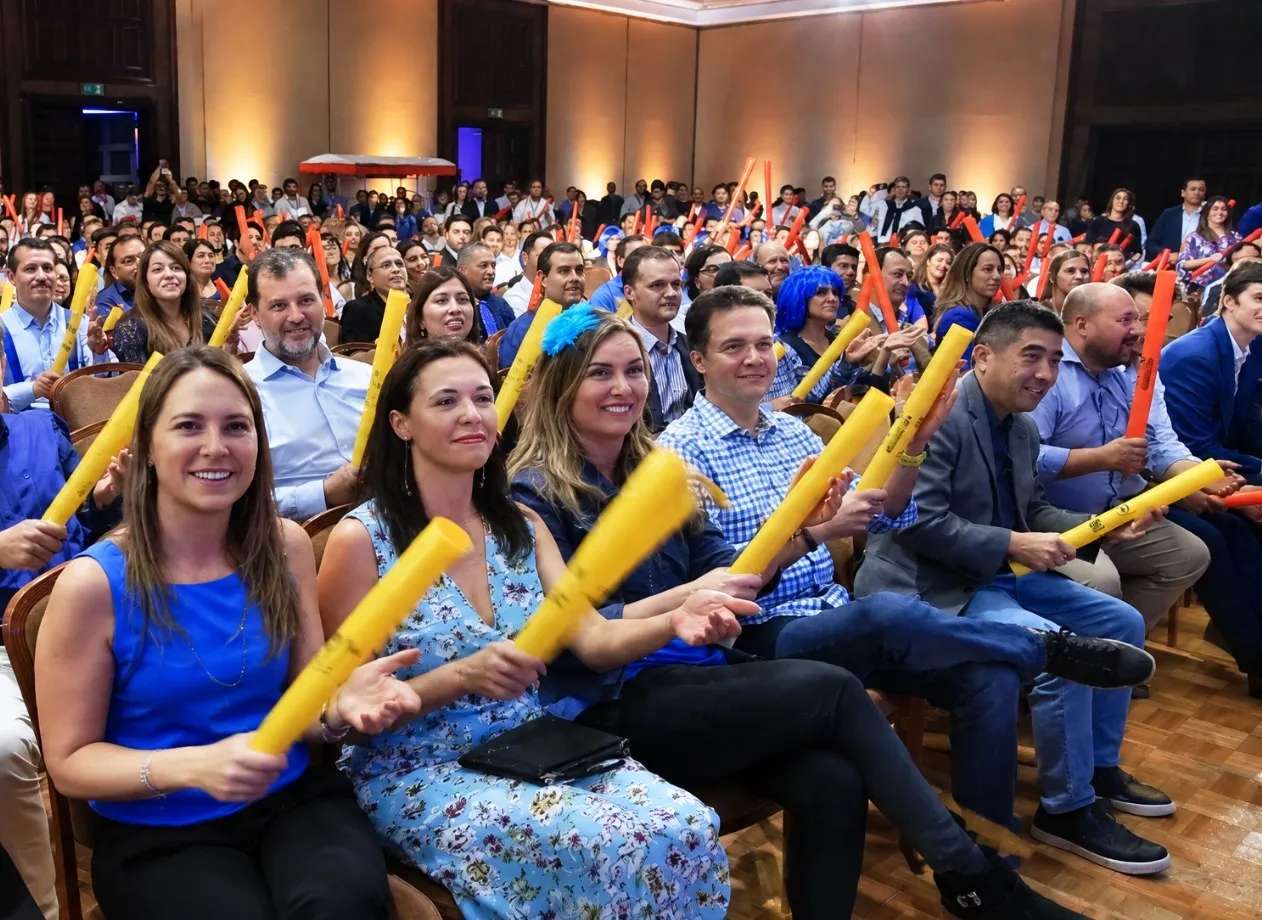 Público sonriente en un salón, levantando tubos de colores amarillos durante una experiencia participativa.