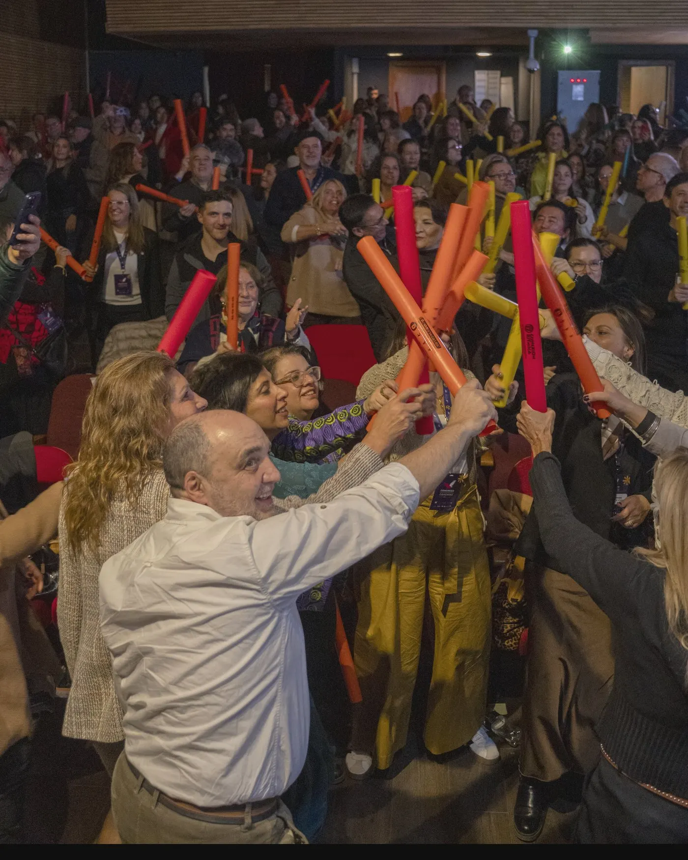 Público en un teatro levantando tubos de colores y sonriendo durante una experiencia participativa.