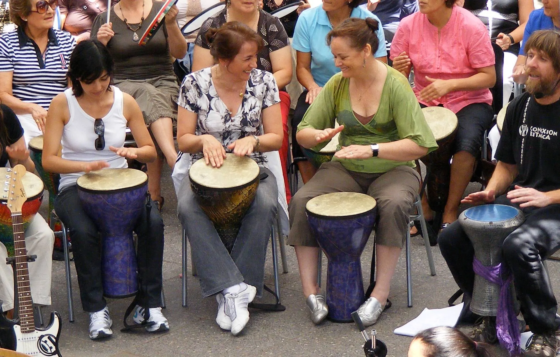 Dos mujeres sonríen y se miran mientras tocan tambores djembé en un círculo al aire libre; a la derecha, un facilitador con la polera de Conexión Rítmica también toca.