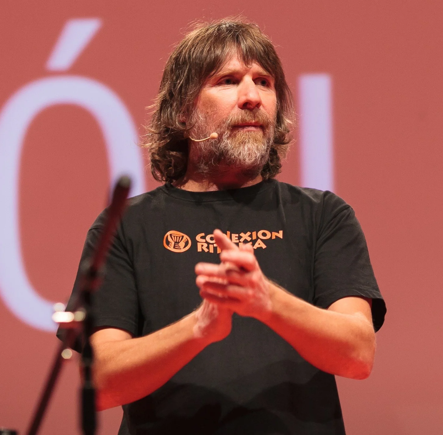 Marcos Ribas en el escenario, con micrófono de diadema y la polera de Conexión Rítmica, aplaudiendo con las manos juntas frente a una pantalla roja.