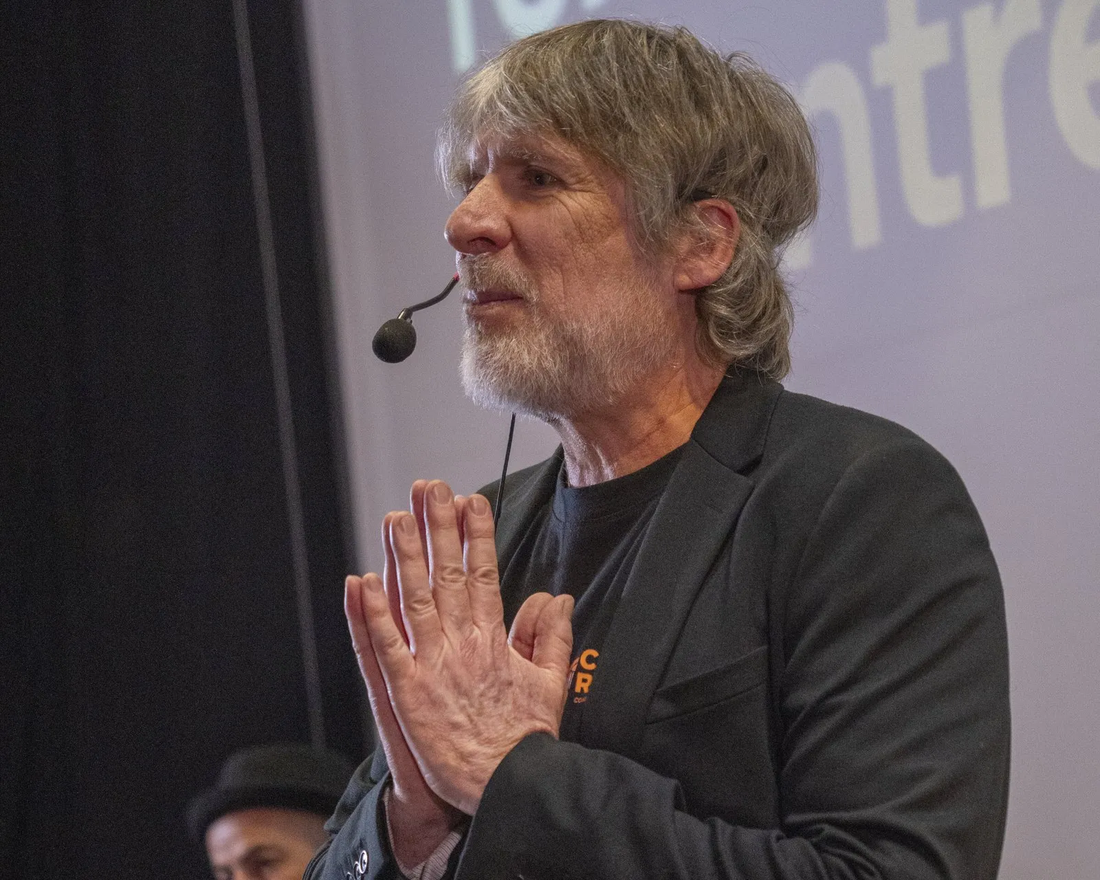 Marcos Ribas con las manos juntas y micrófono de diadema, hablando en un escenario durante una conferencia.