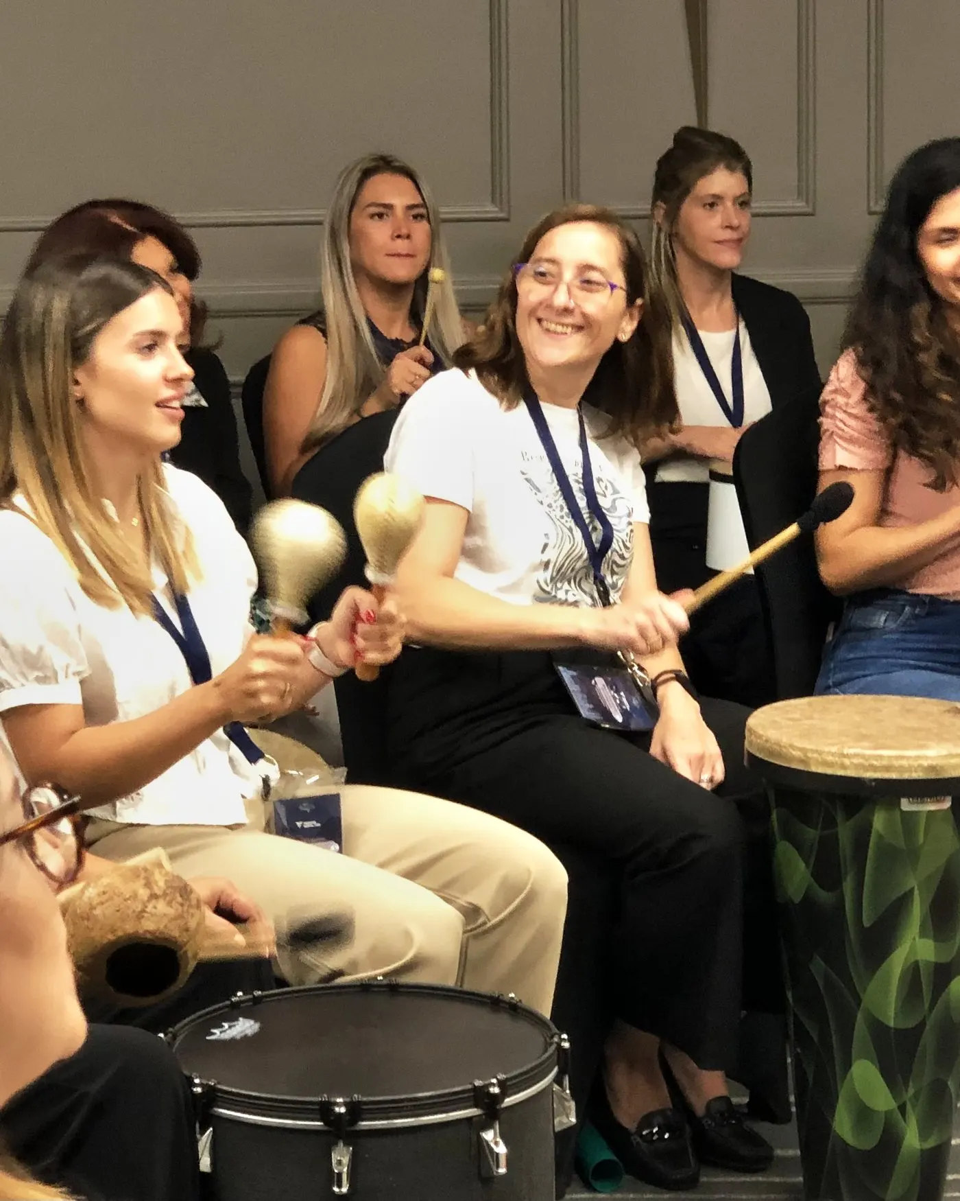 Mujeres sonriendo y tocando maracas y tambores durante una experiencia de Conexión Rítmica en una sala de reuniones.